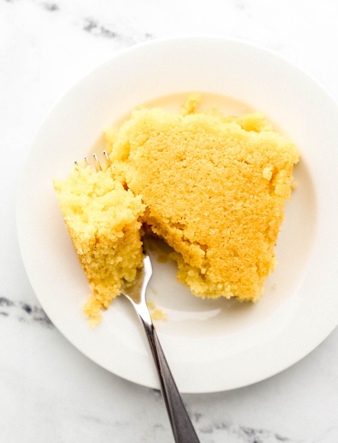 Close up overhead view of piece of cornbread on white plate with fork in it.