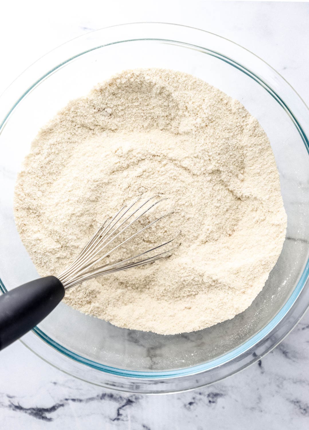 Overhead view of dry ingredients combined in large glass mixing bowl with a whisk.