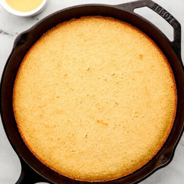 Overhead view of finished honey cornbread in cast iron skillet by small bowl of honey butter sauce.