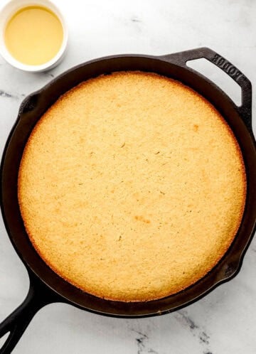 Overhead view of finished honey cornbread in cast iron skillet by small bowl of honey butter sauce.