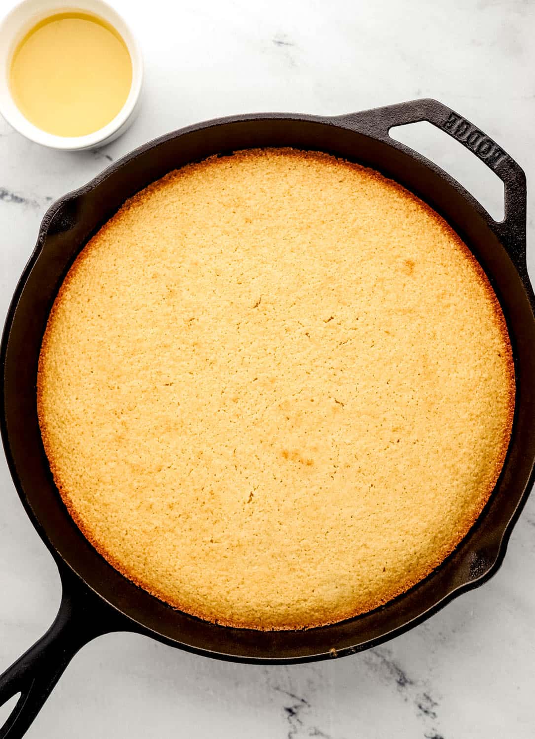 Baked cornbread in cast iron skillet on marble surface by small bowl of honey butter sauce.
