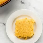 Overhead view of piece of cornbread on white plate with the rest of the cornbread in the background.