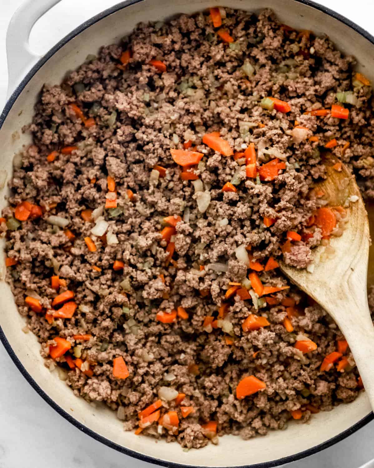 Ground beef added to sauteed vegetables in large white braiser with wooden spoon. 