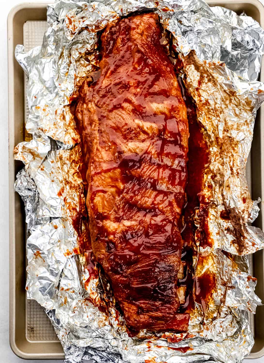 Overhead view of bbq sauce added to ribs on top of foil on sheet pan. 