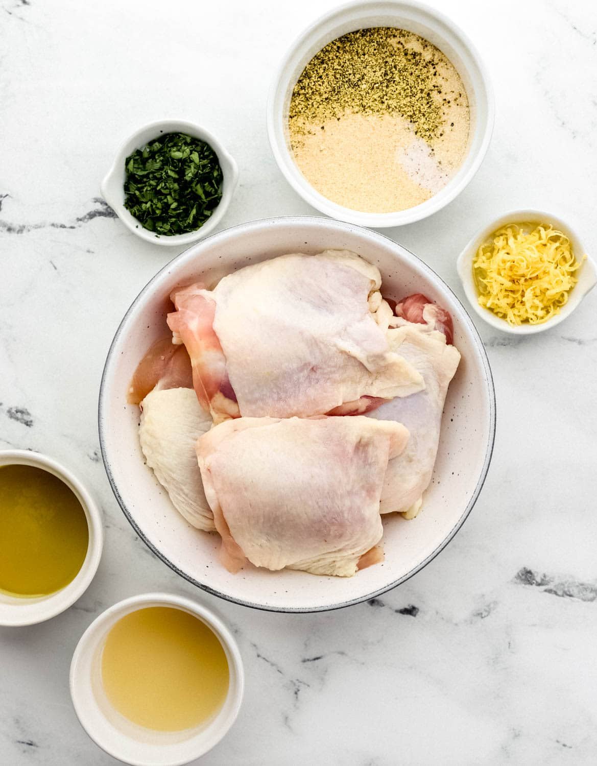 Overhead view of ingredients needed to make chicken in separate bowls on marble surface. 