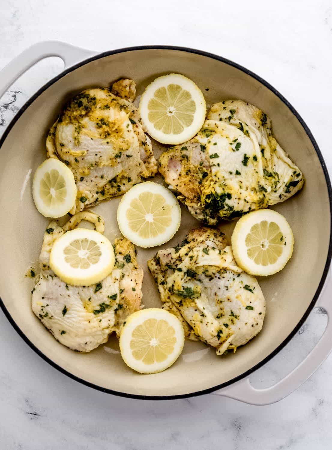 Overhead view of marinated chicken thighs in large braiser pan topped with lemon slices. 
