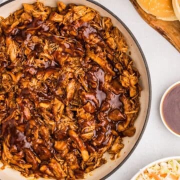 Overhead view of pulled chicken topped with sauce in dutch oven by more sauce, buns, and coleslaw.