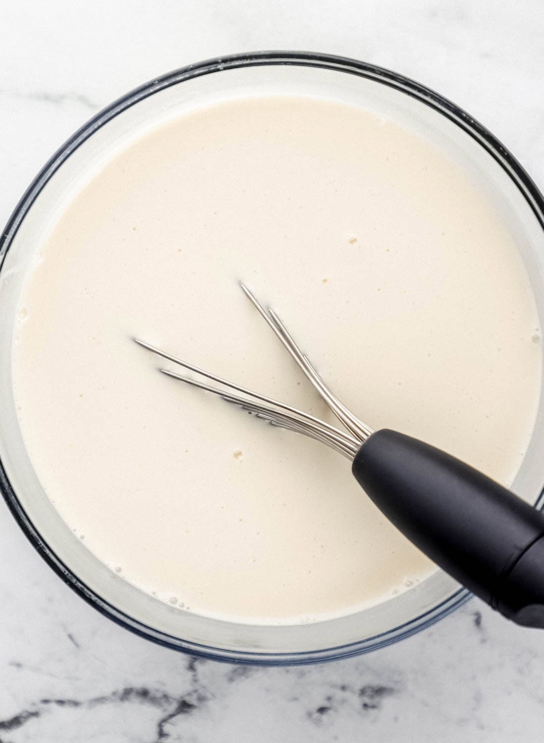 Overhead view of whipped cream ingredients added to large glass mixing bowl with whisk in it.