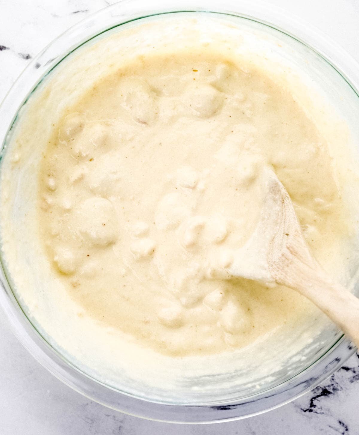 Overhead view of cornbread mixture combined in a large glass mixing bowl with a wooden spoon. 