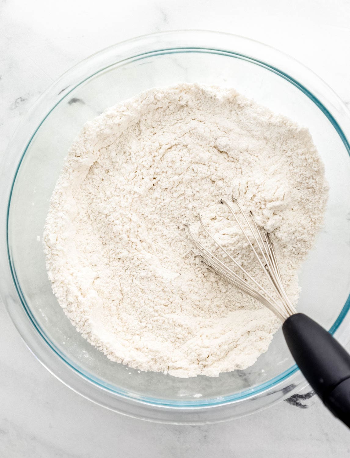 Overhead view of dry ingredients combined in glass mixing bowl with whisk.