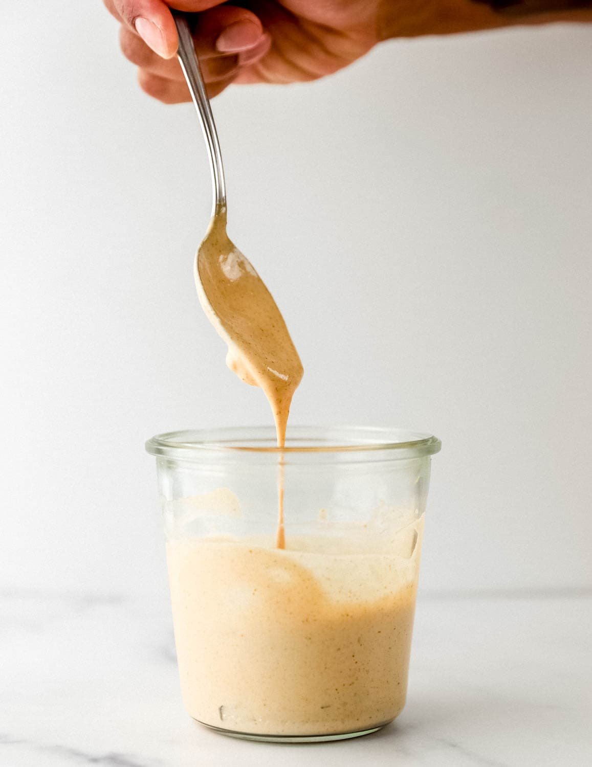 Close up side view of sauce in a jar with hand holding spoon over it. 