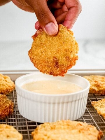 Side view of hand holding fried tomatoes over bowl of sauce.