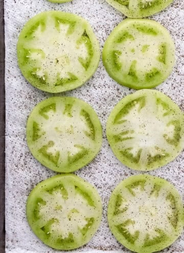 Overhead view of seasoned tomato slices on paper towel lined sheet pan.