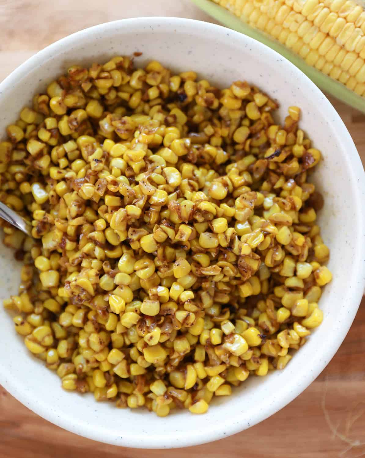 Overhead view of finished corn in a white bowl with a spoon by fresh corn. 