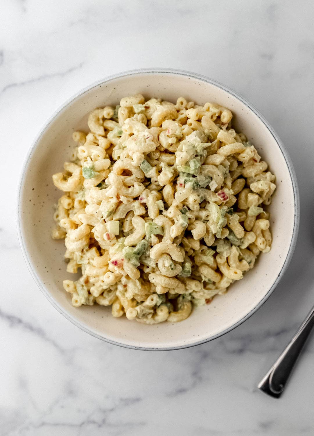Overhead view of finished macaroni salad in white bowl on marble surface. 
