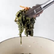 Hand holding tongs with greens on it over pot.