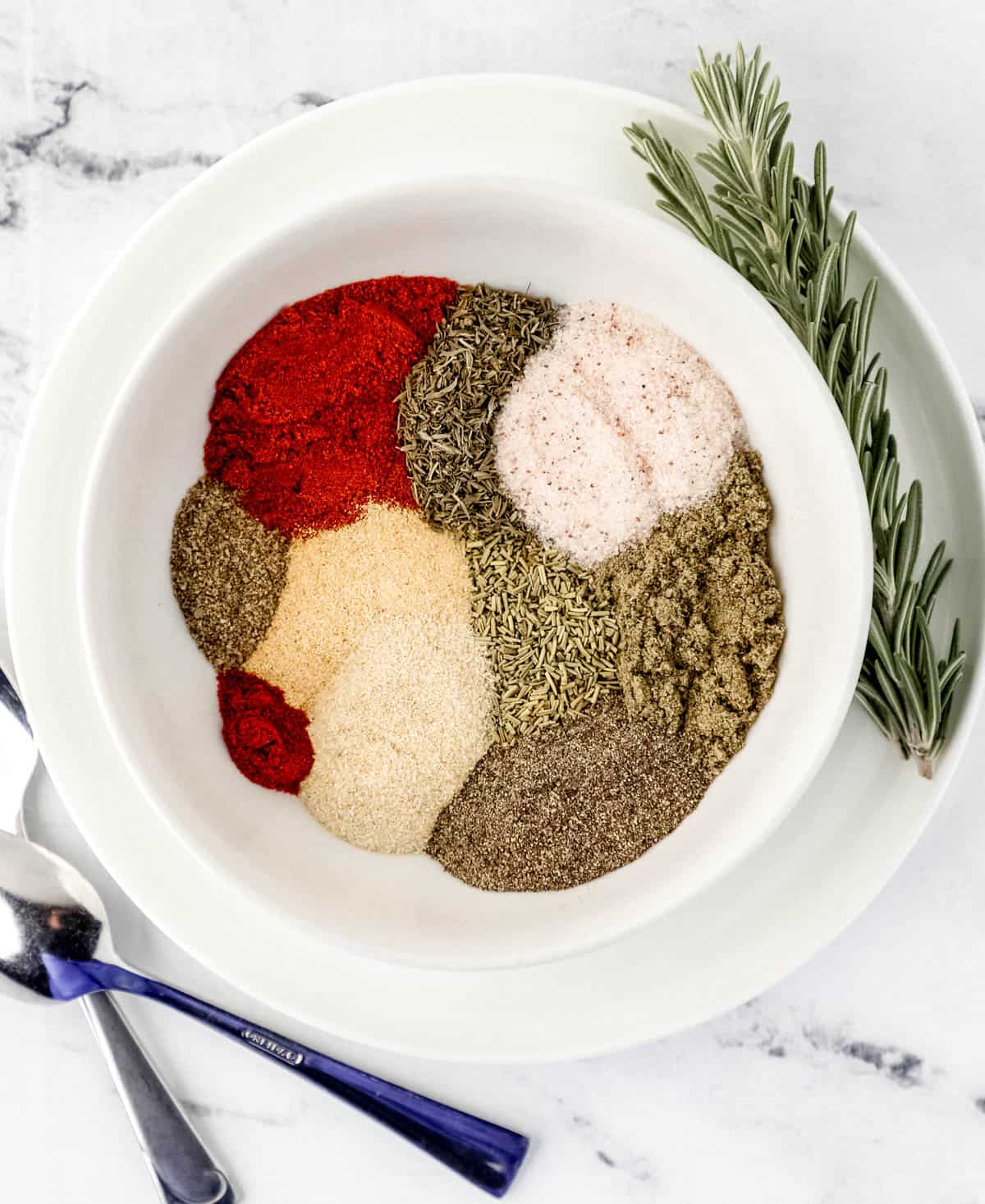 Overhead view of spices in a white bowl by spoons and rosemary. 