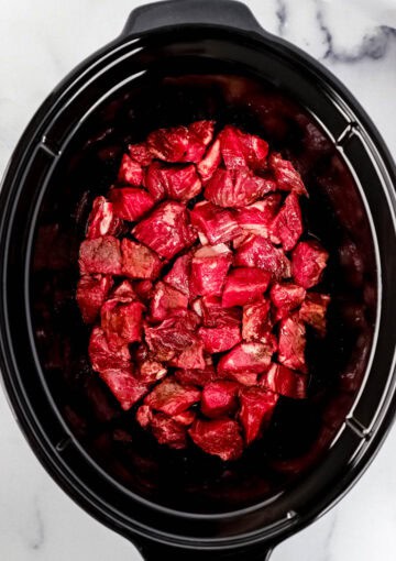 Overhead view of steak bites added to black slow cooker.